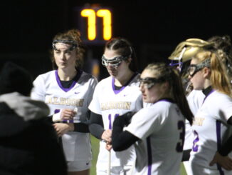 A group of women in white and purple jerseys stand huddled on a field at night, with many players wearing protective gear on their faces. A clock reads ‘77’ in the background in yellow letters.