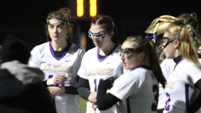 A group of women in white and purple jerseys stand huddled on a field at night, with many players wearing protective gear on their faces. A clock reads ‘77’ in the background in yellow letters.