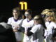 A group of women in white and purple jerseys stand huddled on a field at night, with many players wearing protective gear on their faces. A clock reads ‘77’ in the background in yellow letters.