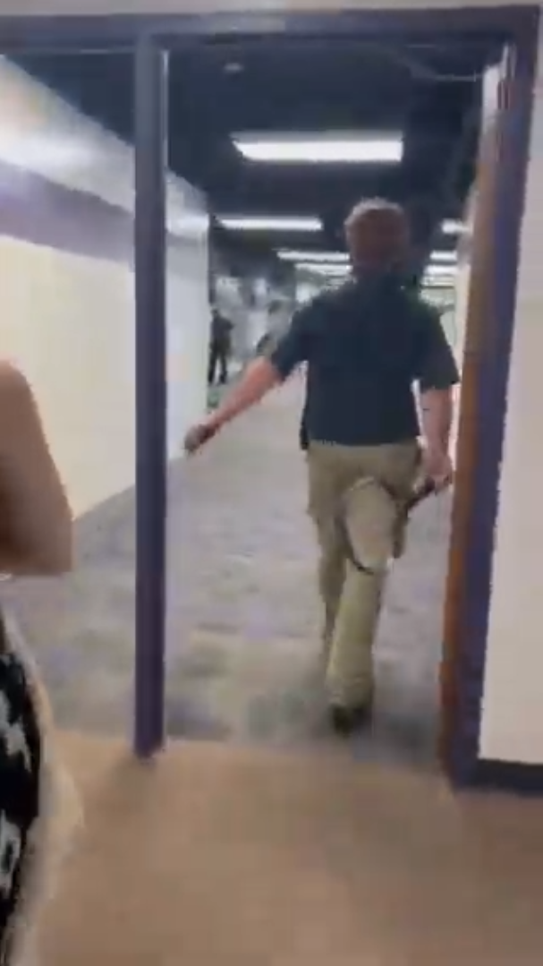 A grainy, low-quality vertical photo of a man from the back wearing a dark polo and khakis walking through a doorway. A second person’s arm is visible at the left edge of the photo. The man swings his arms as he walks, holding a tennis racket in one hand.