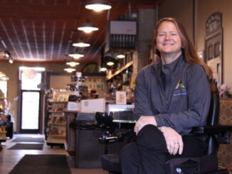 An older woman with long brown hair sits in a black motorized chair, arms crossed and smiling. A store hallway is out of focus behind her, with several bookshelves and a countertop visible, with booths next to her.