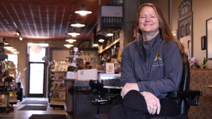 An older woman with long brown hair sits in a black motorized chair, arms crossed and smiling. A store hallway is out of focus behind her, with several bookshelves and a countertop visible, with booths next to her.