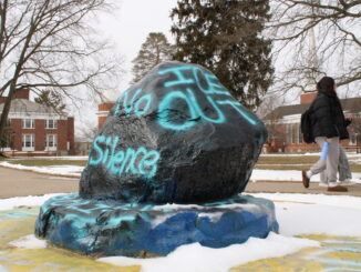 A large rock on a slightly broader stone pedestal sits at the center of the image, painted black with bright, light blue words reading “ICE OUT” and “No Silence” spray-painted on the sides. Snow has accumulated around the base of the rock and on the ground in the background, and two people walk on the sidewalk behind the rock, looking to the right.