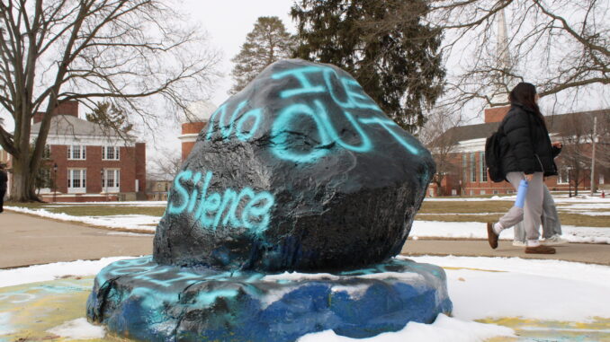 A large rock on a slightly broader stone pedestal sits at the center of the image, painted black with bright, light blue words reading “ICE OUT” and “No Silence” spray-painted on the sides. Snow has accumulated around the base of the rock and on the ground in the background, and two people walk on the sidewalk behind the rock, looking to the right.