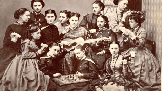 In the foreground of a black and white photo sit four women. The two in the center are playing chess, the one to the right of them is holding binoculars and the one to their left is sitting with a piece of paper in her hands. Behind them are nine other women; the ones in the center are sitting in a row, with the woman in the center playing guitar. The woman to the far left of her is holding some paper, and to the far right, a woman is getting her hair done up with a flower crown whilst reading a piece of paper. All the women are wearing Victorian-era hairstyles and clothing.
