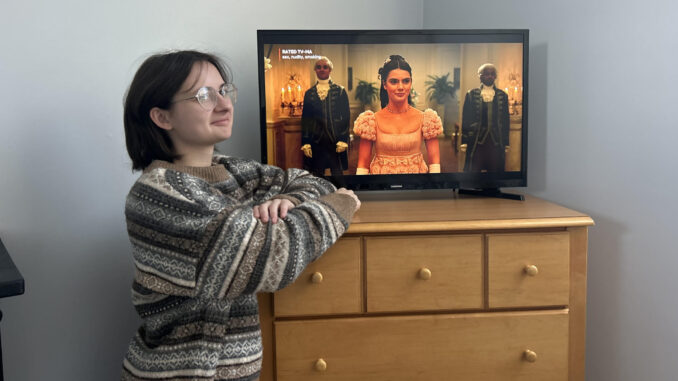 A person with brown hair, glasses and a brown sweater kneels next to a brown dresser with a small TV. The TV shows a character from “Bridgerton” with her hair pinned up and a pink dress with a square neckline and puffy sleeves.