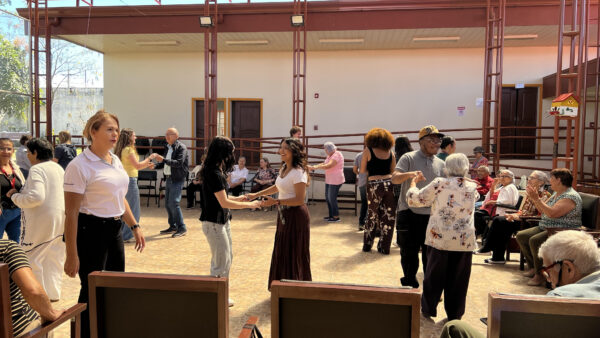 A mixture of young and older individuals are seen either sitting in chairs or dancing. In the foreground, two women are smiling and holding hands. Nearby, a man and an elderly woman hold hands. To the right, a row of seated elderly people are turned towards those standing. Sunlight is seen in the top left corner of the scene.