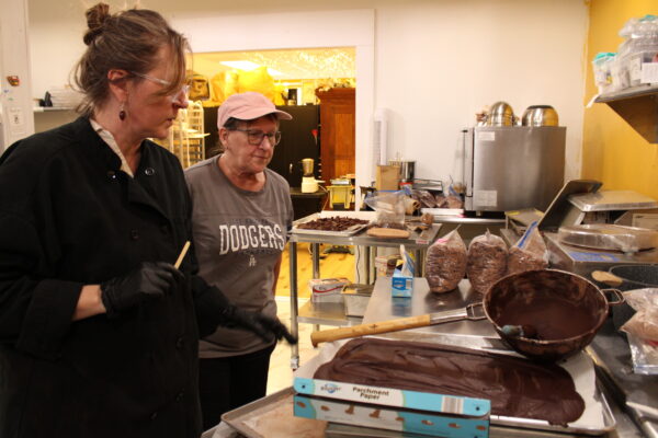 An older woman wearing glasses, an oversized black smock and black latex gloves holds a popsicle stick in one hand while gesturing with her other hand toward a stainless-steel countertop covered in a tray of cooling liquid chocolate, a bowl and several bags of finished chocolate. Next to and behind her, an older woman in a pink baseball cap and glasses watches. They are standing in a cluttered but clean and well-lit kitchen.