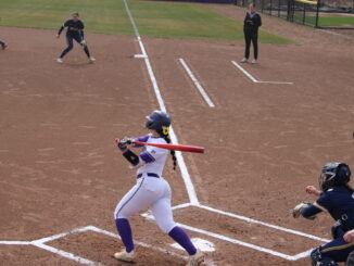 A young woman in a white and purple uniform is captured right after swinging a bright orange baseball bat. She is on a dusty baseball field, standing over the white home plate, in front of two other people in dark blue and black uniforms.
