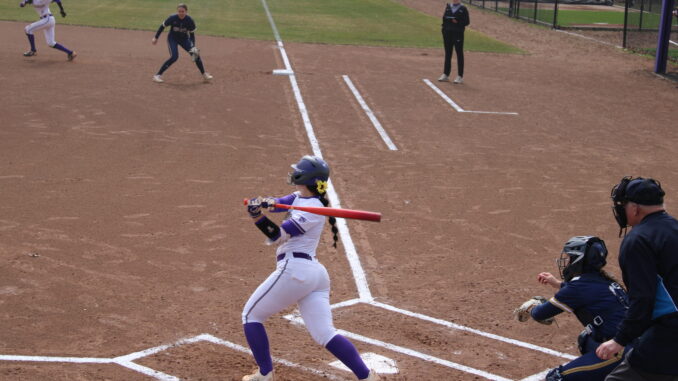 A young woman in a white and purple uniform is captured right after swinging a bright orange baseball bat. She is on a dusty baseball field, standing over the white home plate, in front of two other people in dark blue and black uniforms.