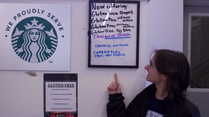 A young person looks and smiles at a hanging frame with multiple gluten-free food options written out on it. They stand against the white wall with the hanging frame, a piece of paper that also lists the gluten-free items and a silver and green metal sign that reads “We proudly Serve” followed by the Starbucks logo.