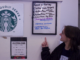 A young person looks and smiles at a hanging frame with multiple gluten-free food options written out on it. They stand against the white wall with the hanging frame, a piece of paper that also lists the gluten-free items and a silver and green metal sign that reads “We proudly Serve” followed by the Starbucks logo.
