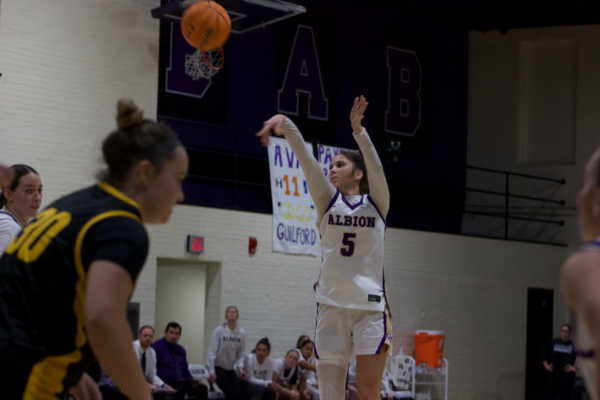 A woman tosses a basketball through the air. She is in a white, purple and gold uniform. Behind her is her team sitting to the side, a white brick wall, senior banners for two of the players and a purple wall with the letters “BAB.”