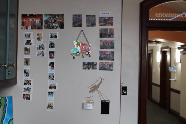 A collection of Polaroids and larger printed-out photos is displayed on a cream-white wall. The photos are arranged in 5 columns, with the leftmost 3 containing the widest variety of photos. Hanging under the 4th and 5th columns is a metal art piece.