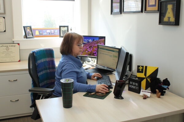 A woman looks at a computer screen in an office space, one hand on a computer mouse and the other resting on the keyboard. She is sitting in an office chair at a desk with various objects such as a purple and gold Albion College shield, business cards and a plush squirrel.