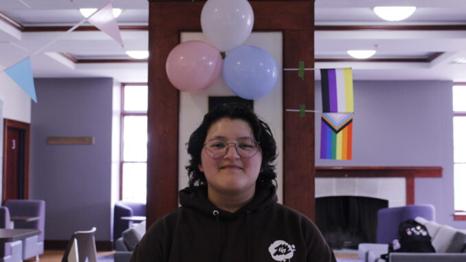 A young person stands in front of a pillar decorated with pink, blue and white balloons representing the transgender flag. Taped to the edge of the pillar is a rainbow flag along with a black, purple, white and yellow non-binary flag. Hanging from the other side of the pillar are more pink, white and blue decorations.