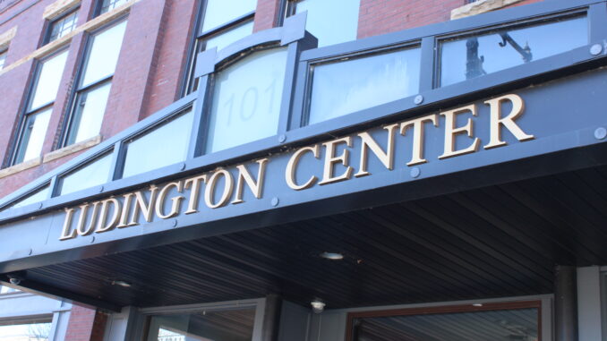 A photograph of the outside of the Ludington Center in downtown Albion. There is a large black sign centered in the photograph with gold letters spelling out ‘Ludington Center,’ along with the building number 101 above it in transparent text.