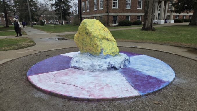 A landscape photo of a large painted rock, with a tiered concrete platform and sidewalk surrounding it. The rock and the platform are painted in white, yellow and purple, though pink and light blue paint is visible beneath. People walk by on the sidewalk in the background.