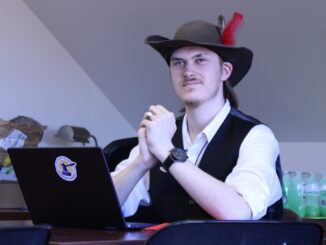 A young adult wearing a hat with a red feather and a vest sits at a desk with a laptop, hands clasped, in a room with a slanted ceiling.