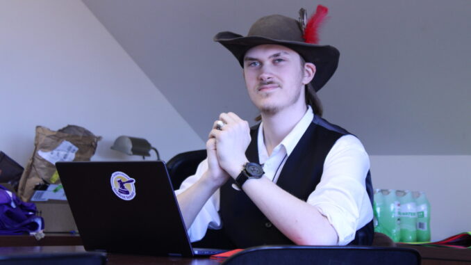 A young adult wearing a hat with a red feather and a vest sits at a desk with a laptop, hands clasped, in a room with a slanted ceiling.