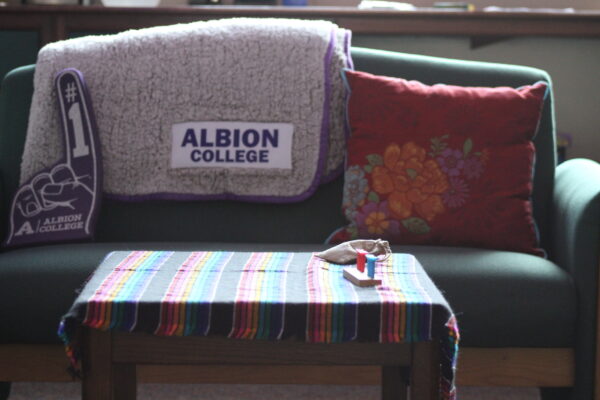 A forest green couch is displayed with a grey blanket draped over it with the words “ALBION COLLEGE” stitched into it. To the left of the blanket is a dark red pillow with a yellow chrysanthemum embroidered onto it. The couch also has a foam finger on it with the words “#1 ALBION COLLEGE.”